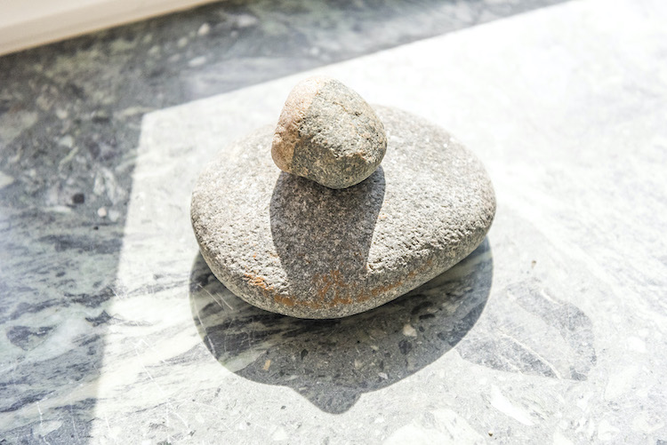 Grau gesprenkelter Steinboden, auf den die Sonne scheint. Auf dem Boden ein bräunlicher Stein, auf dem ein kleiner ebenfalls bräunlicher Stein liegt.