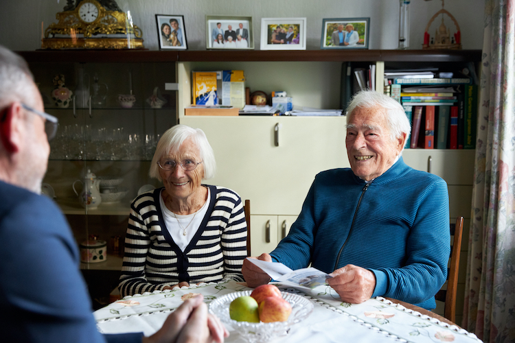 Zwei alte Männer und eine alte Frau sitzen im Wohnzimmer an einem Esstisch und lächeln.